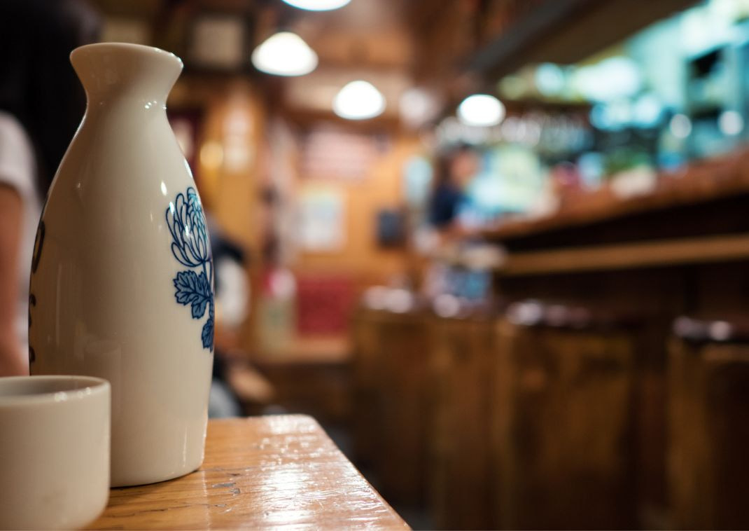 Un flacon de saké en céramique et une tasse sur une table en bois à l’intérieur d’un bar ou restaurant japonais chaleureux.