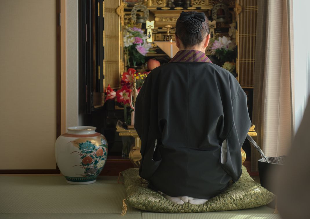 Une personne en kimono formel agenouillée sur un coussin, priant devant un autel familial bouddhiste décoré de fleurs et d’offrandes.