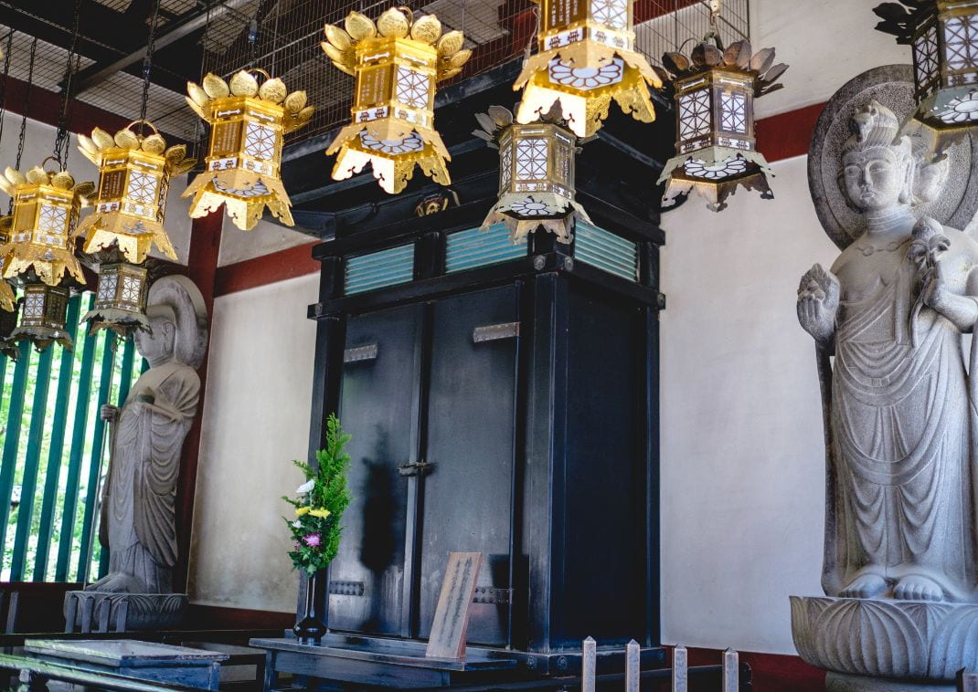 Des lanternes dorées suspendues au-dessus d’un grand autel noir encadré par des statues bouddhistes en pierre dans un temple japonais.