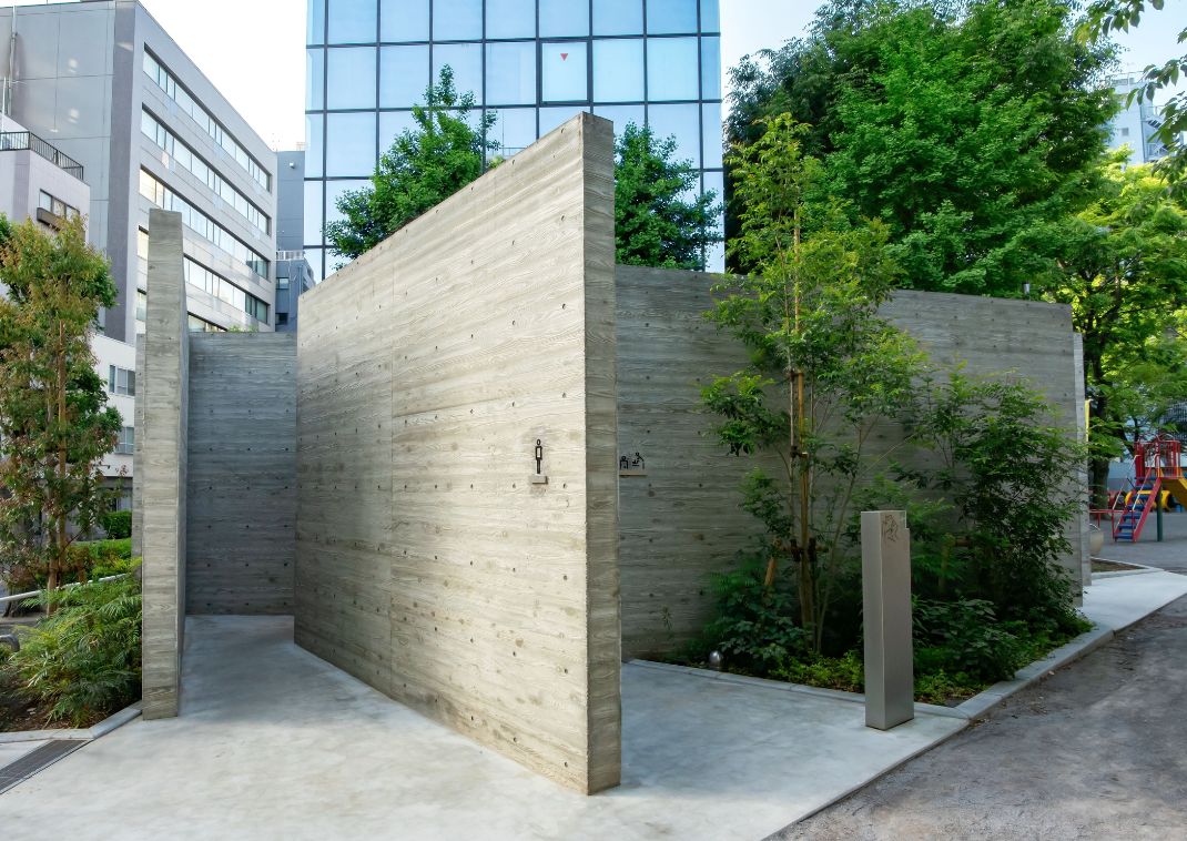 Des toilettes publiques en béton brut au design minimaliste, entourées de plantes et d’arbres, dans un environnement urbain.