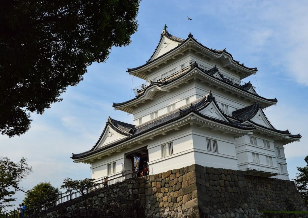 Château japonais traditionnel blanc d’Odawara, perché sur une colline de pierres, avec des toits noirs et des arbres en arrière-plan sous un ciel clair.