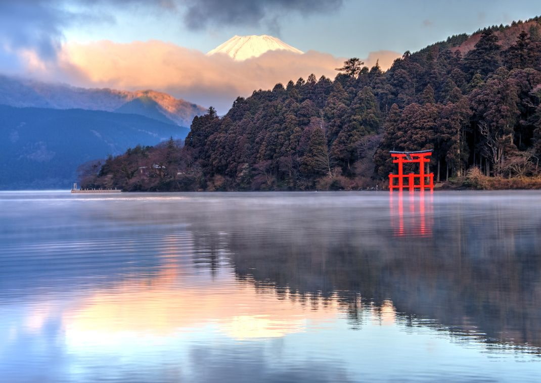 Vue panoramique du torii rouge du sanctuaire Hakone-jinja émergeant de la forêt sur la rive du lac Ashi, avec le mont Fuji enneigé en arrière-plan et des reflets colorés sur l’eau.