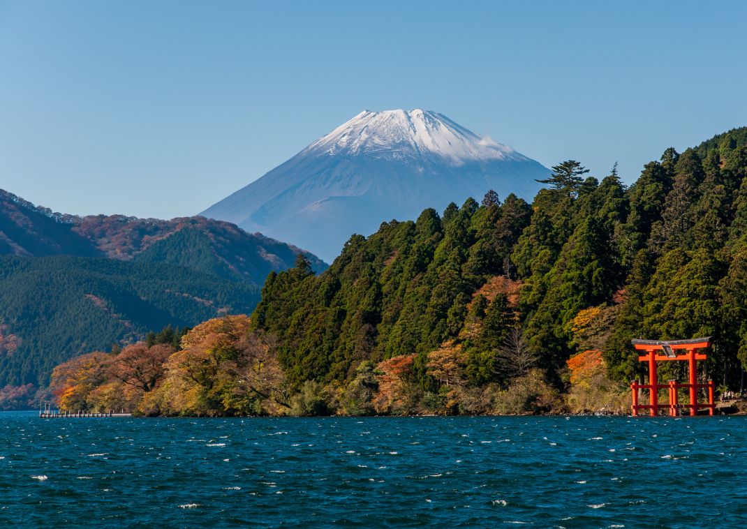 Vue panoramique sur le lac Ashi bordé de forêts et de feuillages d’automne, avec un grand torii rouge du sanctuaire Hakone-jinja et le mont Fuji enneigé en arrière-plan sous un ciel bleu clair