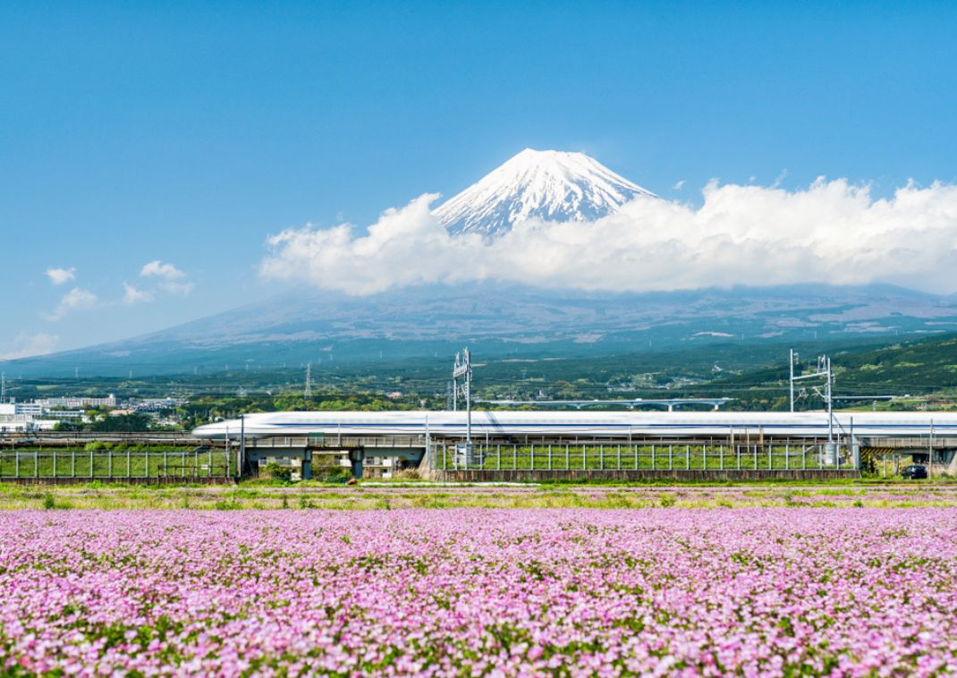 Train à grande vitesse Shinkansen traversant un pont métallique au-dessus d’une rivière, avec le mont Fuji enneigé en arrière-plan sous un ciel bleu clair.