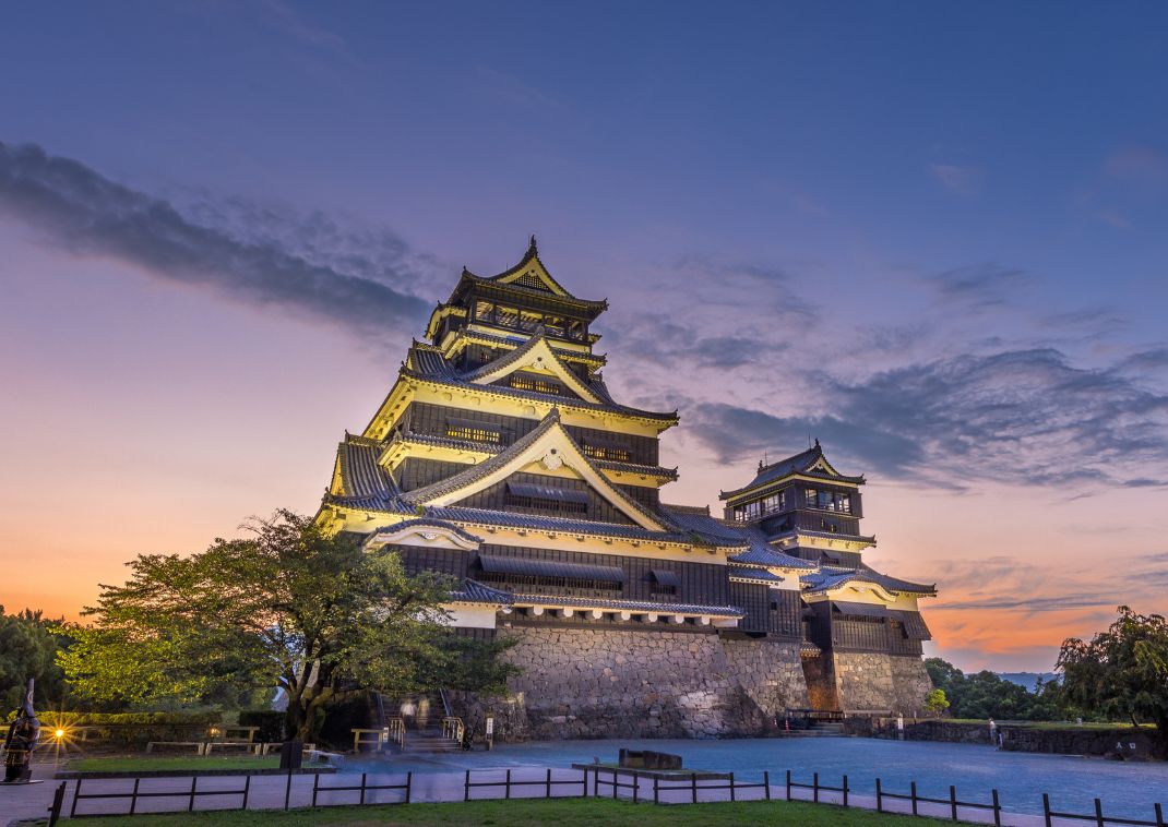 Château japonais traditionnel de Kumamoto illuminé au crépuscule, avec des murs noirs et des toits en tuiles, sous un ciel aux couleurs pastel.