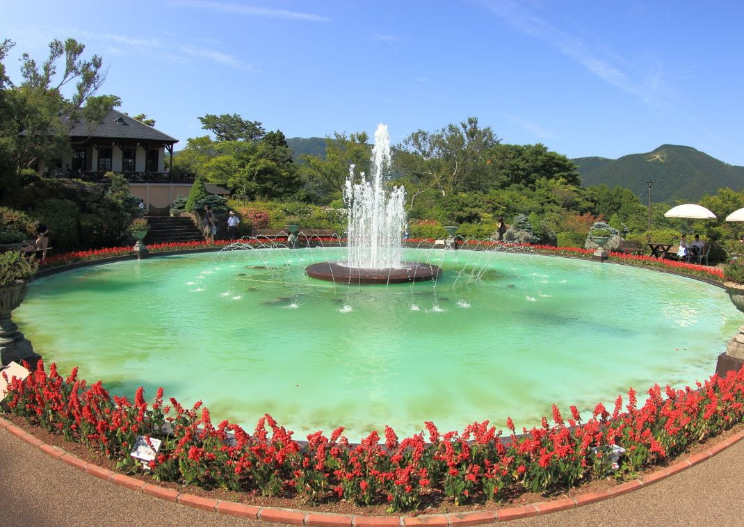 Fontaine circulaire entourée de fleurs rouges dans un jardin paysager luxuriant à Hakone, sous un ciel bleu clair.