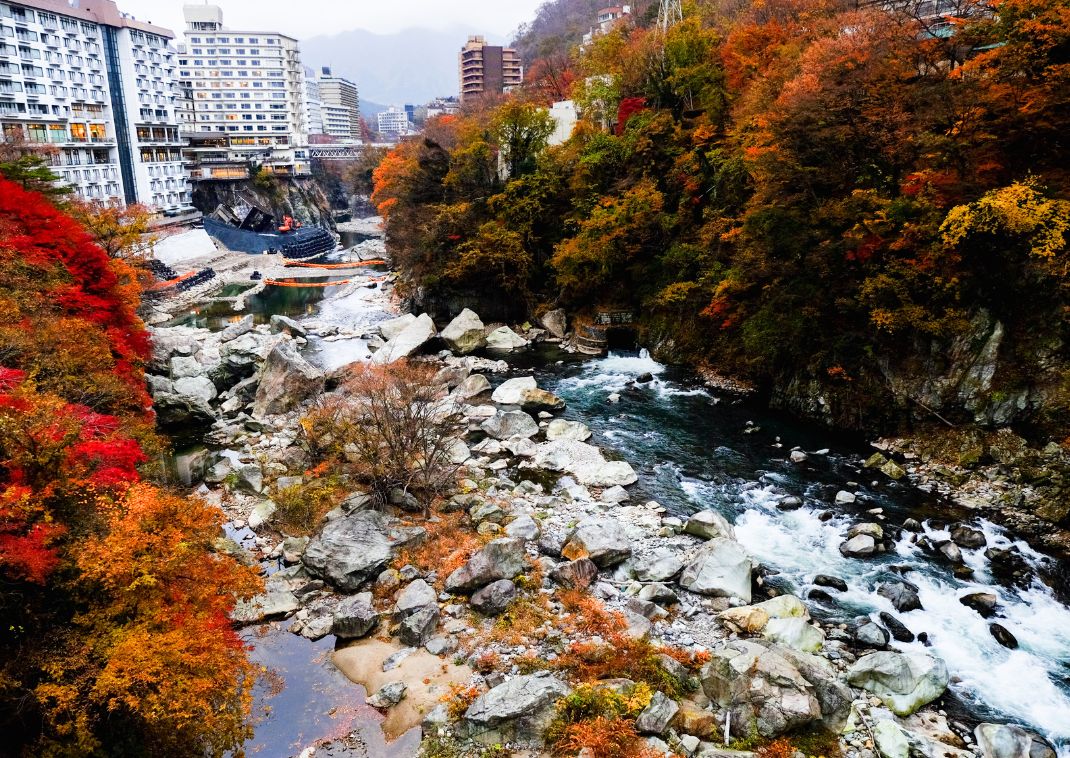 Rivière bordée de rochers et de forêts aux couleurs d’automne traversant une ville thermale japonaise, avec des bâtiments modernes en arrière-plan et des collines boisée