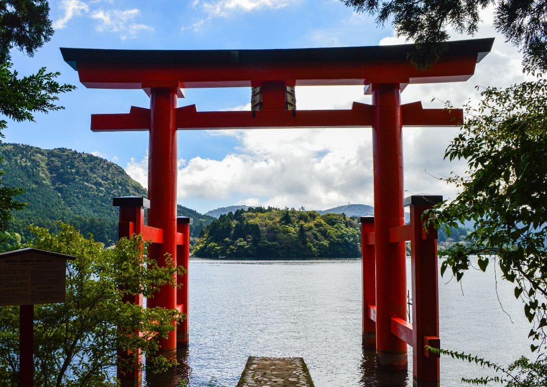 Un grand torii rouge du sanctuaire Hakone-jinja se dresse sur les rives du lac Ashi, encadré par des arbres, avec des montagnes verdoyantes en arrière-plan et le ciel bleu parsemé de nuages.