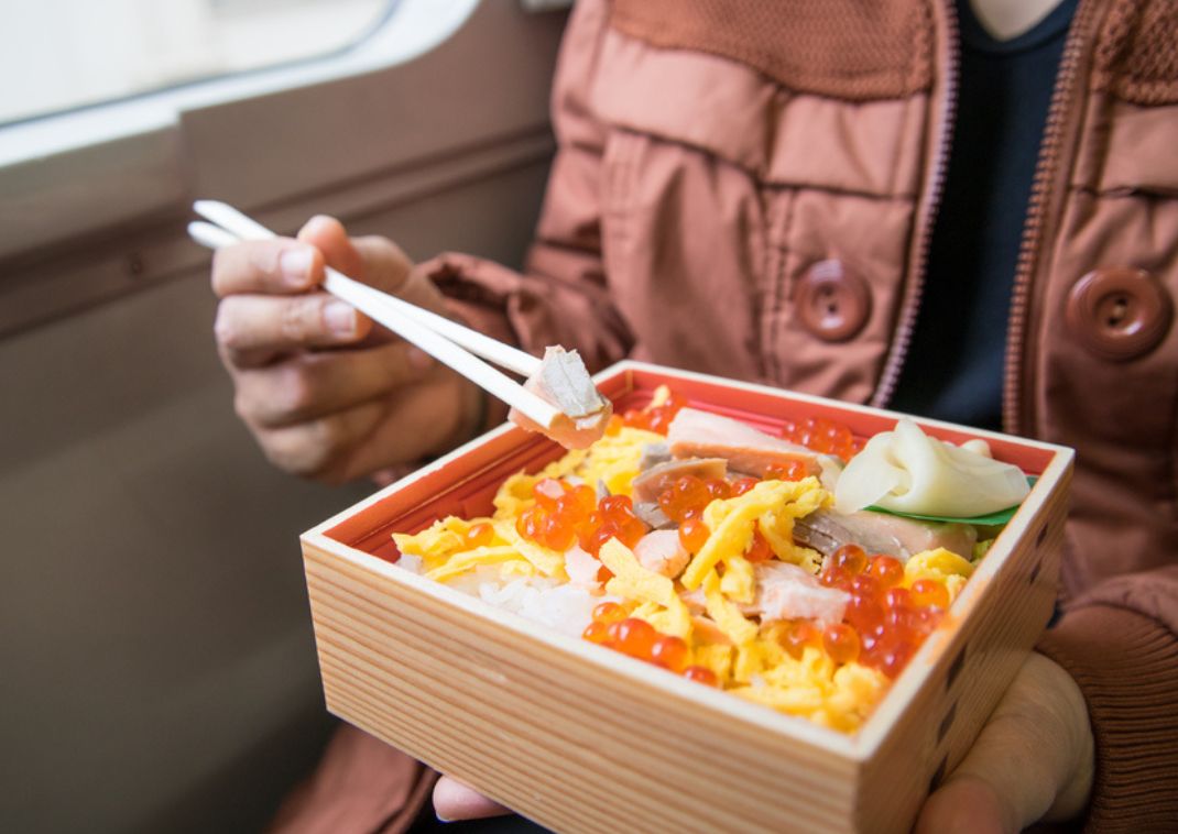 Personne dégustant un bento japonais coloré avec des baguettes, assise près de la fenêtre à bord d’un train.