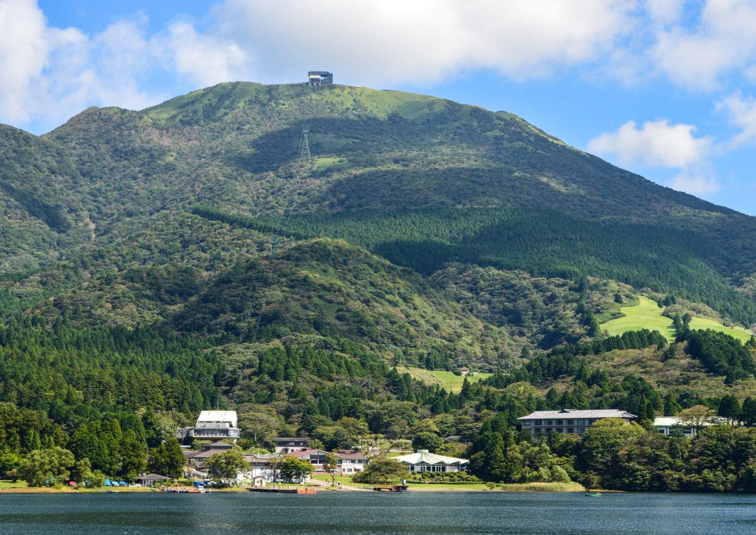 Vue sur le mont Komagatake recouvert de forêts verdoyantes, avec un bâtiment d’observation au sommet et le lac Ashi au premier plan, sous un ciel partiellement nuageux.

