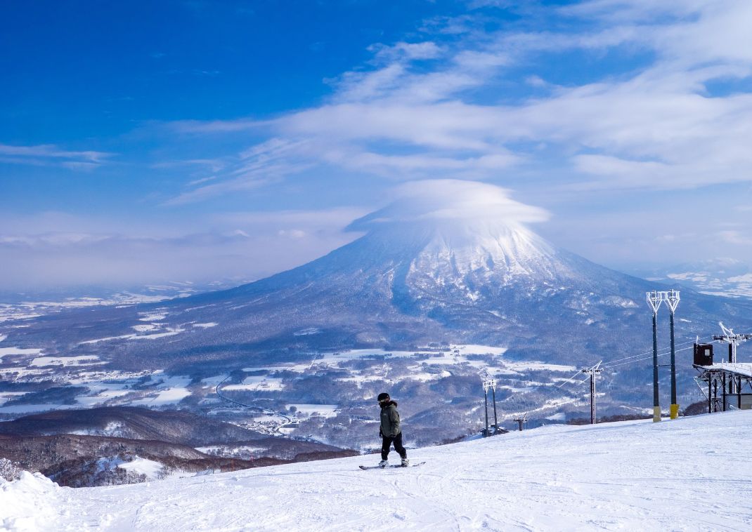 Skieur solitaire descendant une piste enneigée avec le mont Yotei recouvert de neige et coiffé de nuages en arrière-plan sous un ciel bleu éclatant.