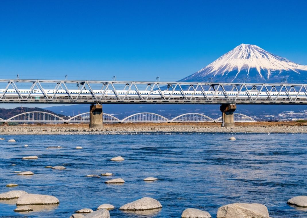 Train à grande vitesse Shinkansen traversant un pont métallique au-dessus d’une rivière, avec le mont Fuji enneigé en arrière-plan sous un ciel bleu clair.