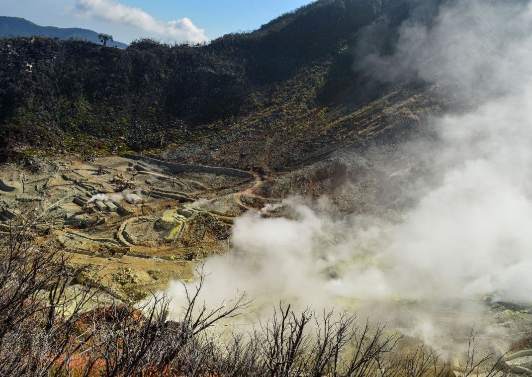 Paysage volcanique de la vallée d’Owakudani à Hakone, avec des fumerolles de soufre s’élevant du sol et des sentiers serpentant à travers la roche, sous un ciel ensoleillé.
