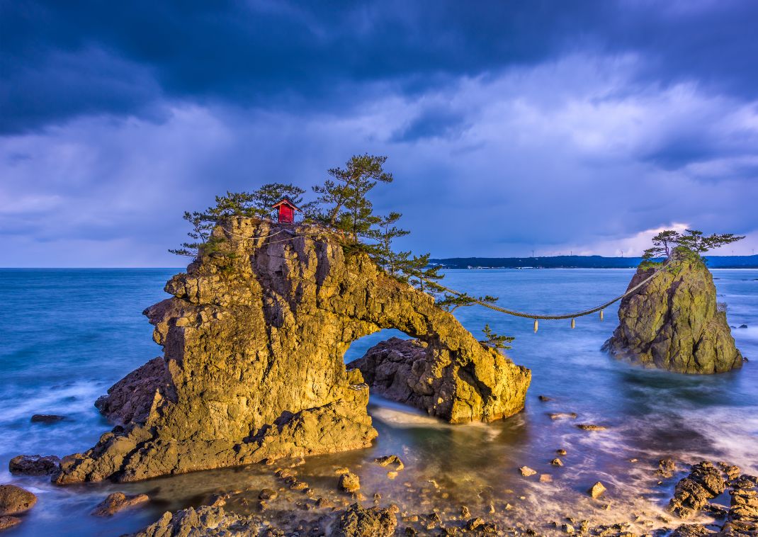 Formation rocheuse spectaculaire en bord de mer à Noto, avec un petit sanctuaire rouge au sommet, des pins et un pont de corde reliant deux rochers, sous un ciel nuageux au crépuscule.
