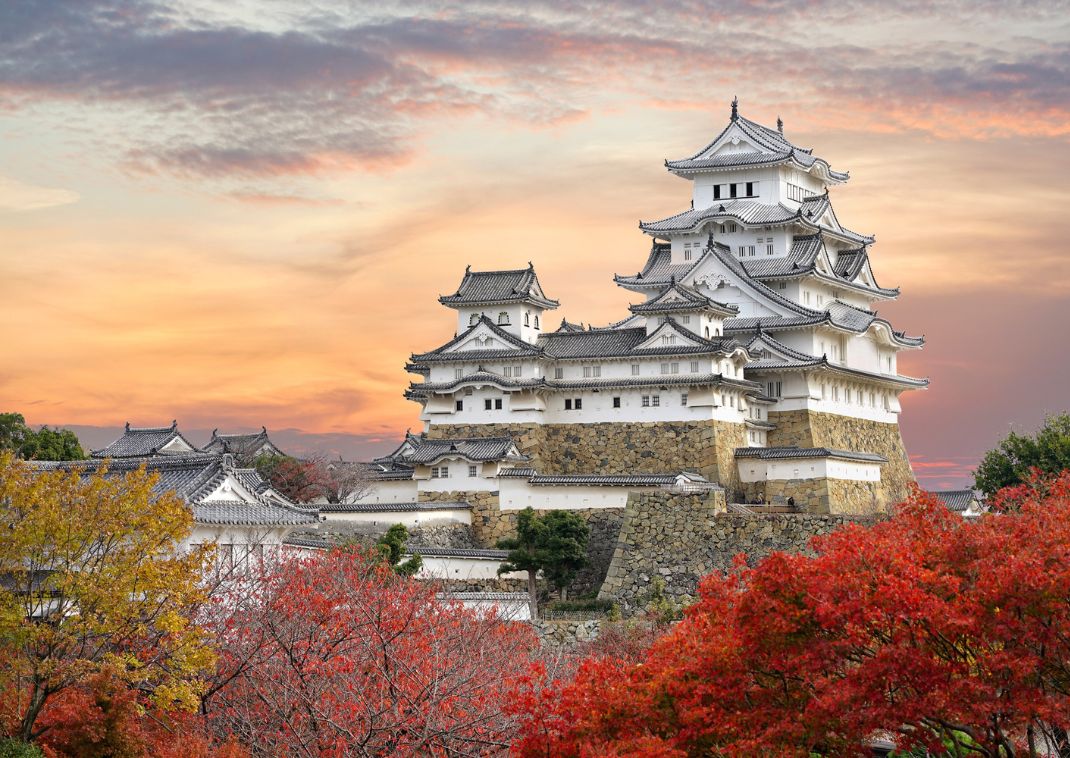 Château japonais traditionnel de Himeji, aux murs blancs et toits en tuiles grises, entouré d’arbres aux feuillages rouges et jaunes en automne, sous un ciel de coucher de soleil aux couleurs pastel.