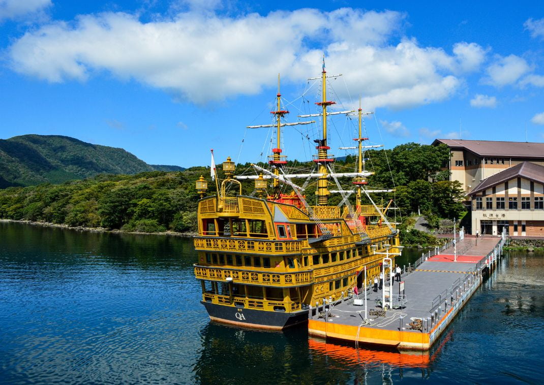 Un bateau de croisière jaune au style de galion amarré sur le lac Ashi, entouré de collines verdoyantes et sous un ciel bleu parsemé de nuages.